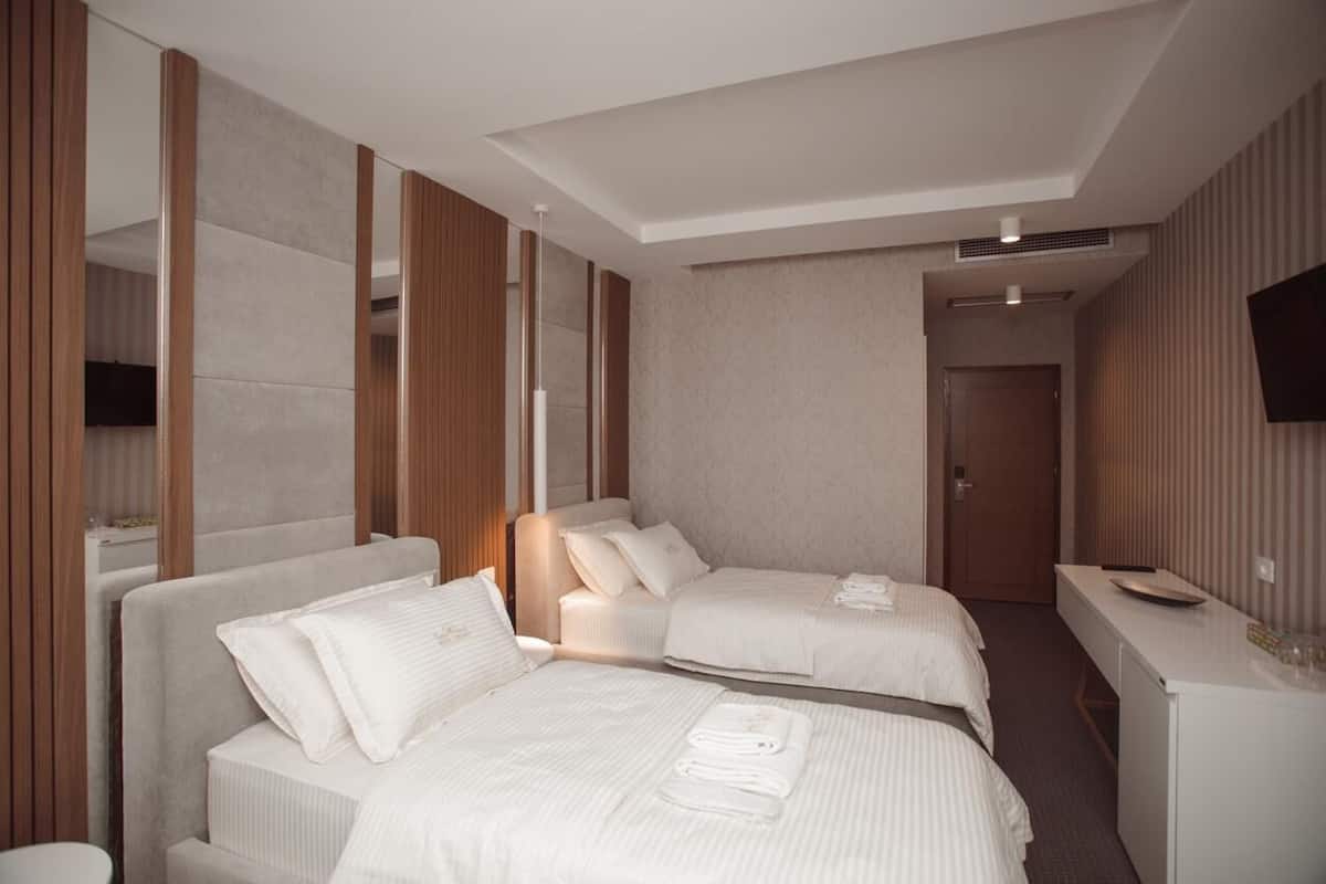 A hotel room with two beds, a desk, a flat-screen TV, and a door leading to another room.