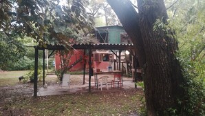 A rustic outdoor seating area with a table and chairs, a small structure with a green roof, and a tree in the foreground. - Cabaña del humedal (Partido de Tigre)