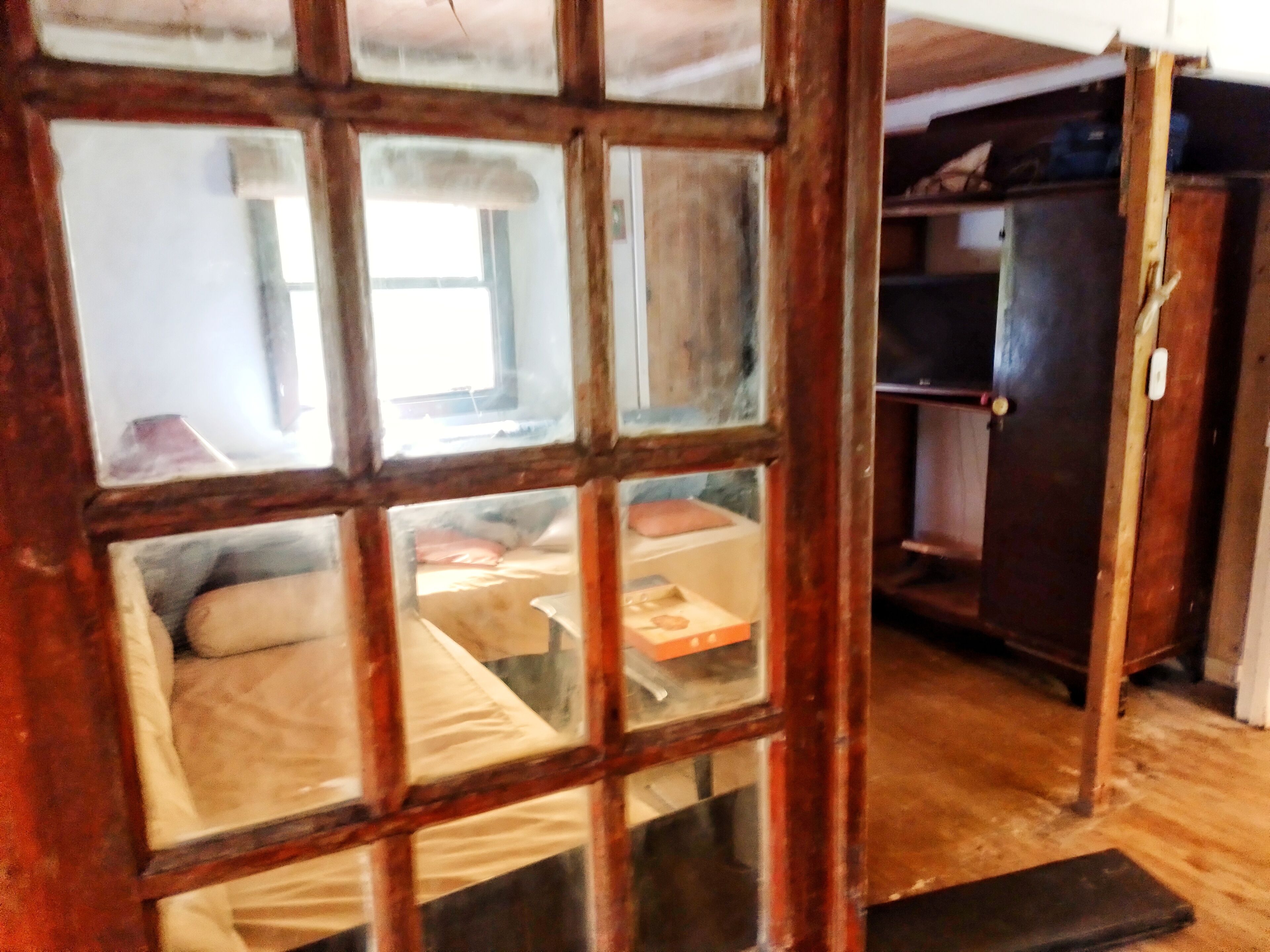 A room with a wooden door, a bed with a white mattress and pillows, a small table with a box, and a wooden cabinet.