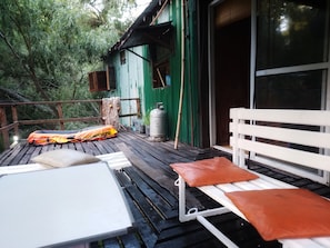 A wooden deck with a white table and orange cushions, a green building with a window, and a white bench. - Cabaña del humedal (Partido de Tigre)
