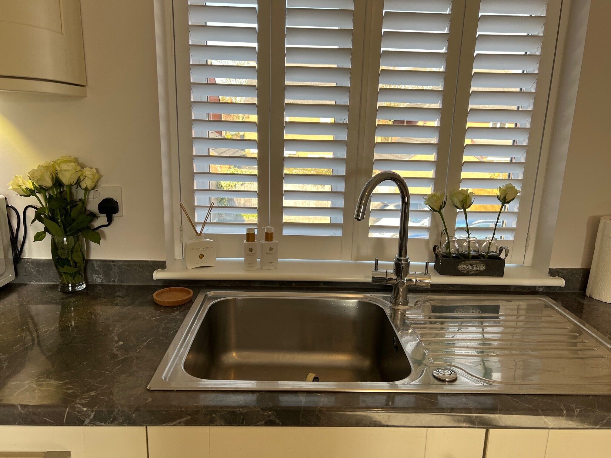 A kitchen with a stainless steel sink, a window with blinds, and a vase with flowers on the counter.