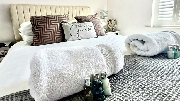 Un lit bien fait, avec un oreiller orné de l’inscription « SOYEZ NOTRE INVITÉ », une serviette enroulée et deux flacons de produits de toilette posés sur une couverture à motifs.