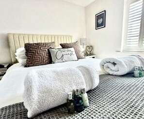 Ein ordentlich bezogenes Bett mit einem Kopfkissen mit der Aufschrift "BE OUR Guest“, einem aufgerollten Handtuch und zwei Flaschen Pflegeprodukten auf einer strukturierten Bettdecke.