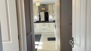 Un couloir mène à une cuisine dotée de meubles blancs, d’un plan de travail noir et d’un luminaire suspendu.