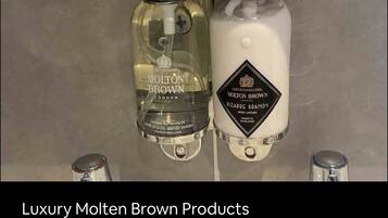 Två produkter från Molten Brown, en flytande handtvål och en kroppslotion, visas upp på en vägg täckt med kakel.