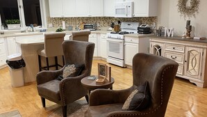 A modern kitchen with white cabinets, a microwave, and a stove. There are two brown leather chairs with cushions, a small round table, and a decorative plant.