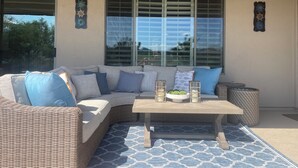 A sunlit patio with a wicker sofa, blue cushions, a wooden table, and decorative wall art.