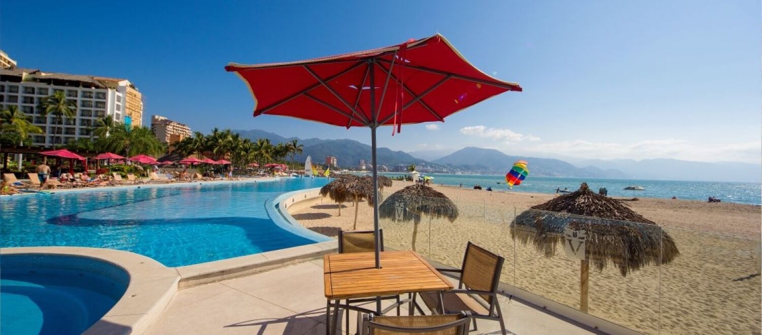 A resort pool with a red umbrella, beach chairs, and a dining table.