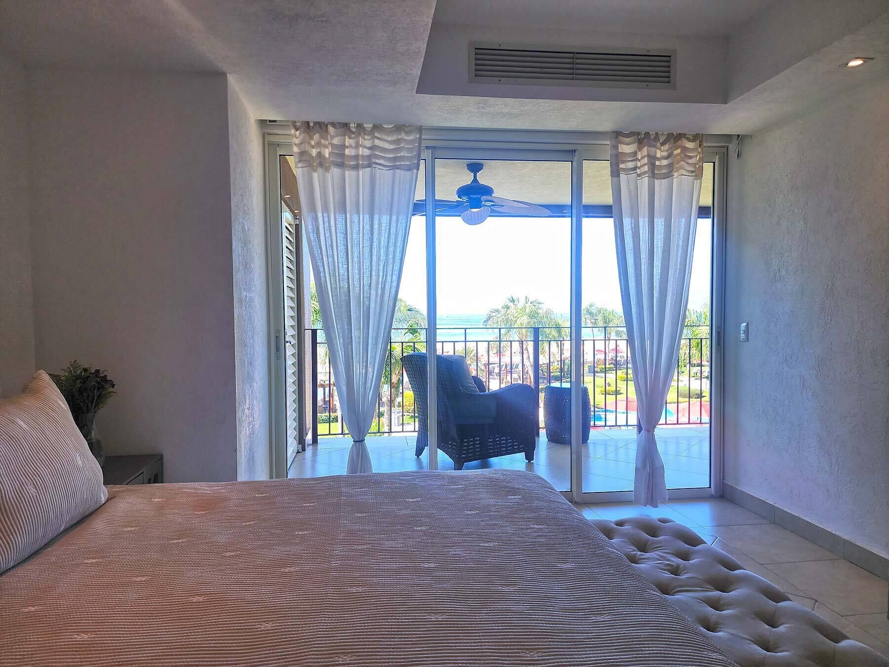 A bedroom with a large bed, a ceiling fan, and a view of a balcony with a chair and palm trees.