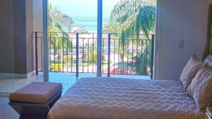 A bedroom with a large bed, a bench, and a view of a pool and palm trees through a sliding glass door.