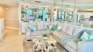 A living room with a sectional sofa, a glass coffee table, and a kitchen in the background. - Float The Stress Away & Enjoy The Lazy River. Pet Friendly Condo On 5th Floor (Orange Beach)