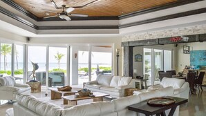 A spacious living room with white leather sofas, a wooden ceiling, and large windows offering a view of the ocean. - GoldenEye Estates!- GoldenEye + Palm Villas (Freeport)
