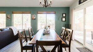 Un comedor con una mesa y sillas de madera, una araña y ventanas amplias.