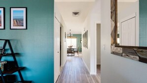 Un pasillo con paredes azul verdoso, suelo de madera y una estantería metálica negra con libros. En la distancia, se ve un comedor con una mesa y sillas.