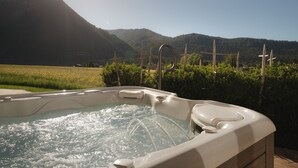 A hot tub with a wooden exterior, surrounded by a fence and greenery, with mountains in the background.