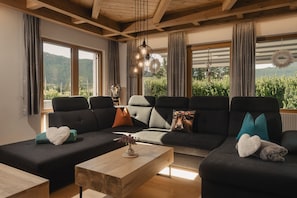 A modern living room with a sectional sofa, a wooden coffee table, and large windows offering a view of greenery.