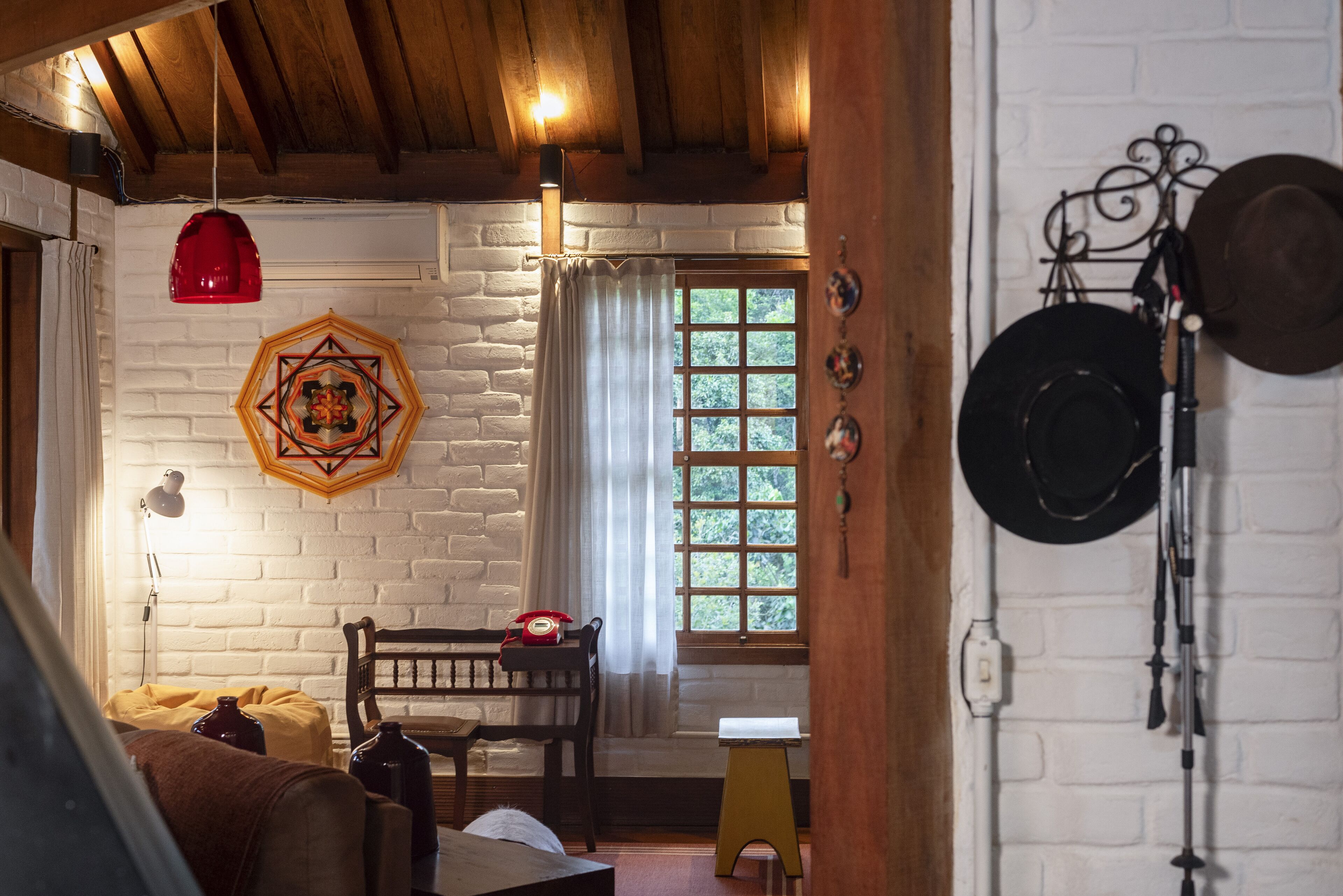 Una habitación rústica con techo de madera, pared de ladrillo blanco, un banco de madera, una mesita y una ventana con cortinas.