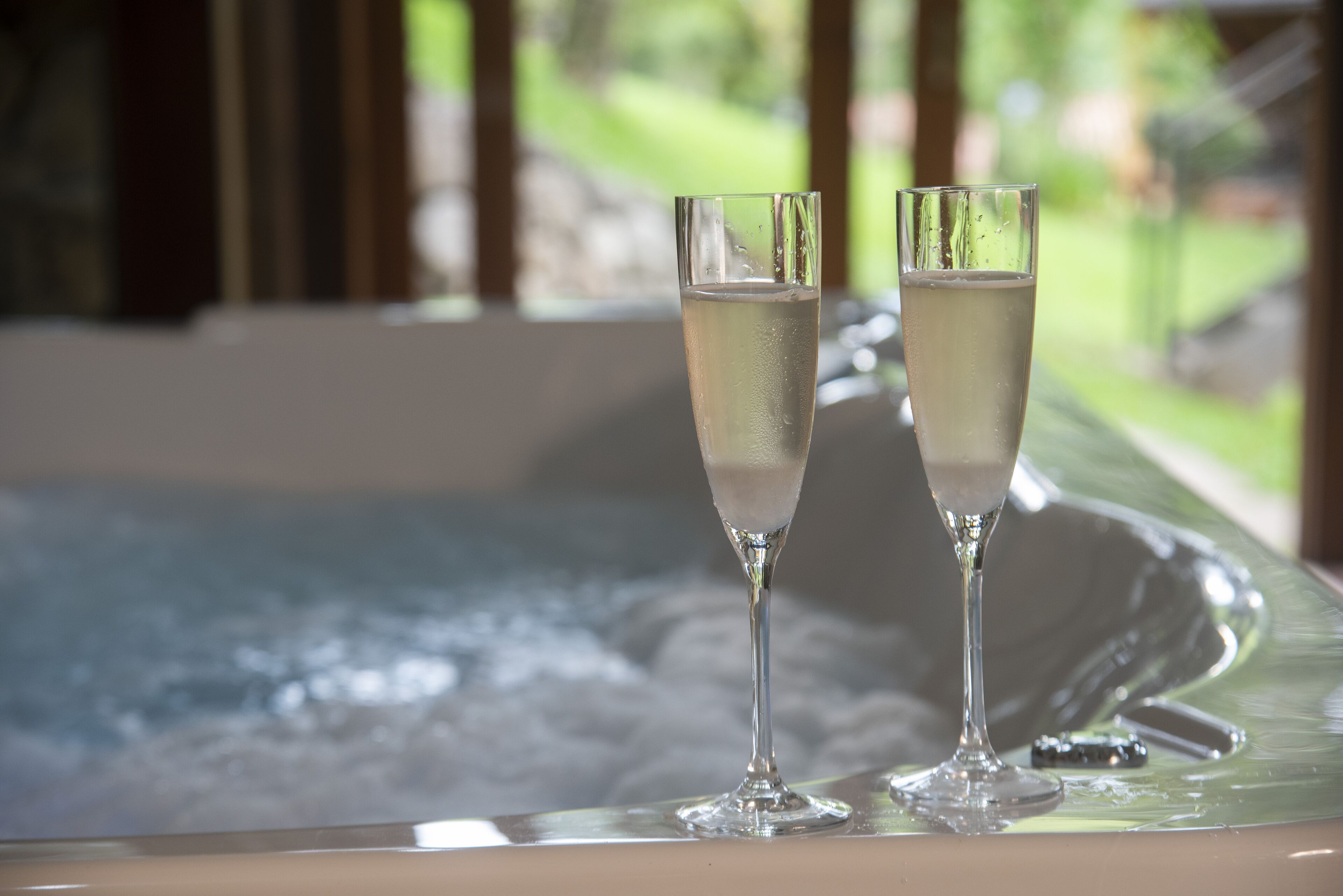 Two champagne flutes on a reflective surface, likely a table or counter, with a blurred outdoor view in the background.