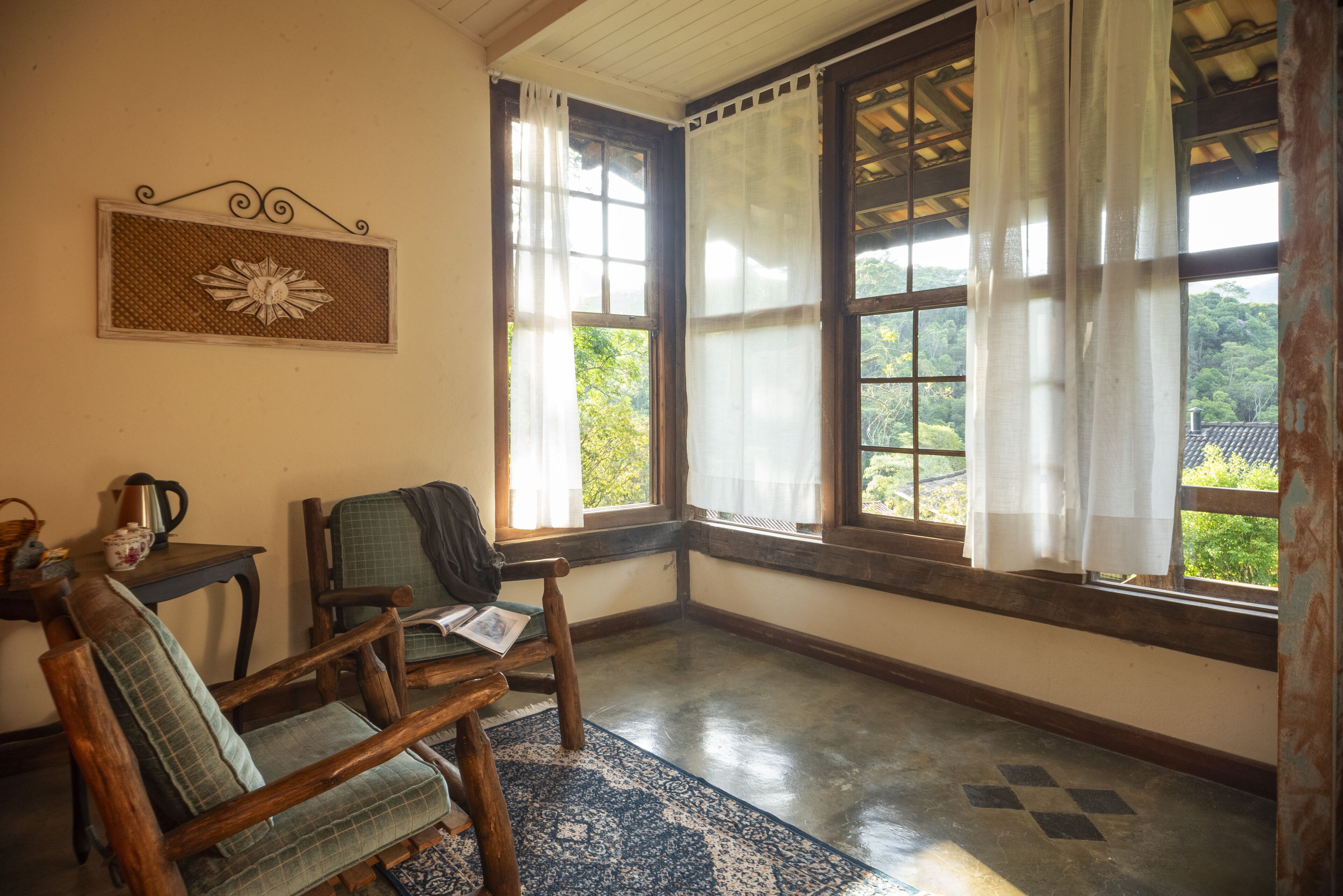 A room with a wooden bench, a small table with a kettle, and a view of greenery through large windows.