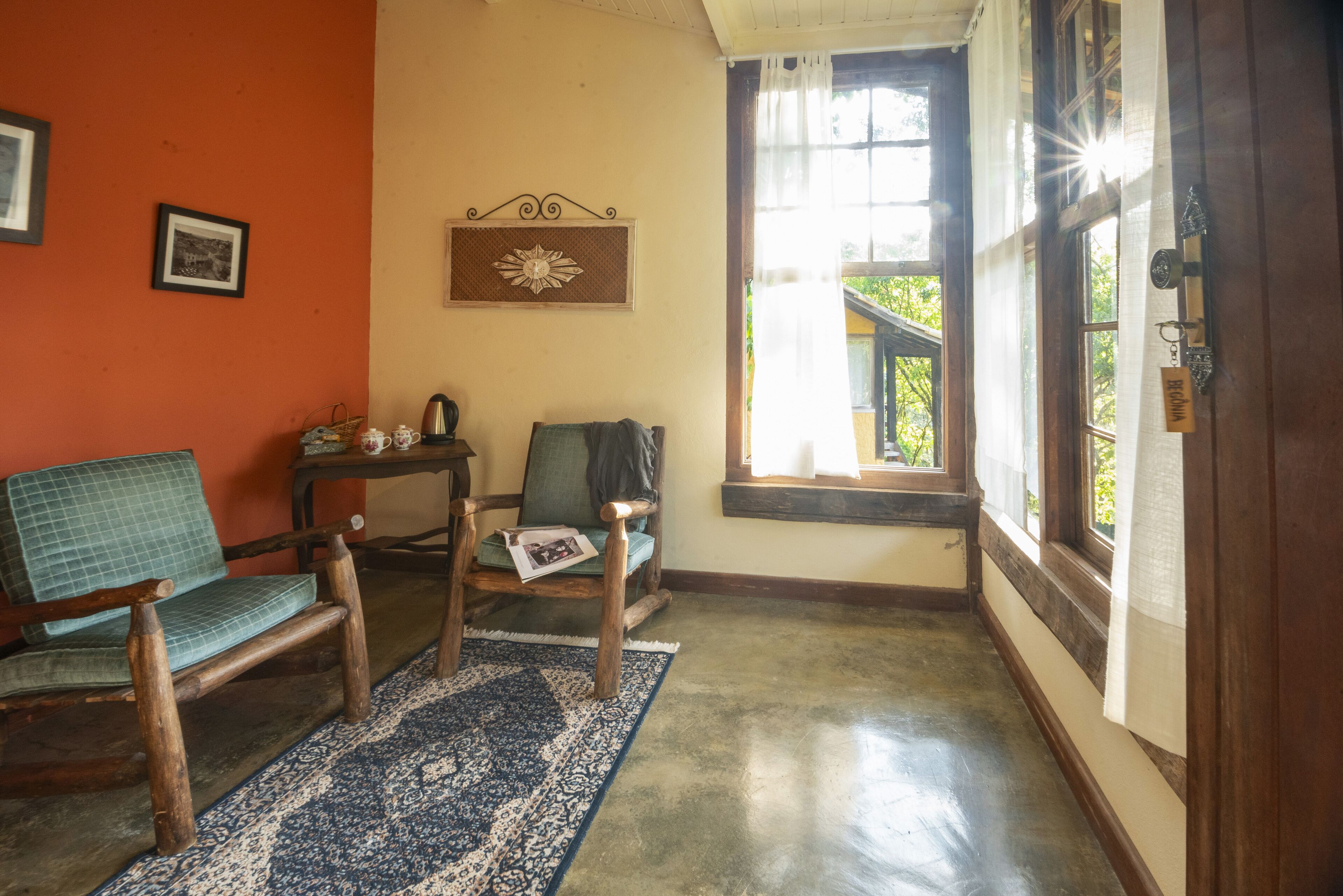 Una habitación con piso de madera, un sofá tapizado en verde, una silla de madera, una mesita con tetera y un ventanal con vistas a la vegetación.