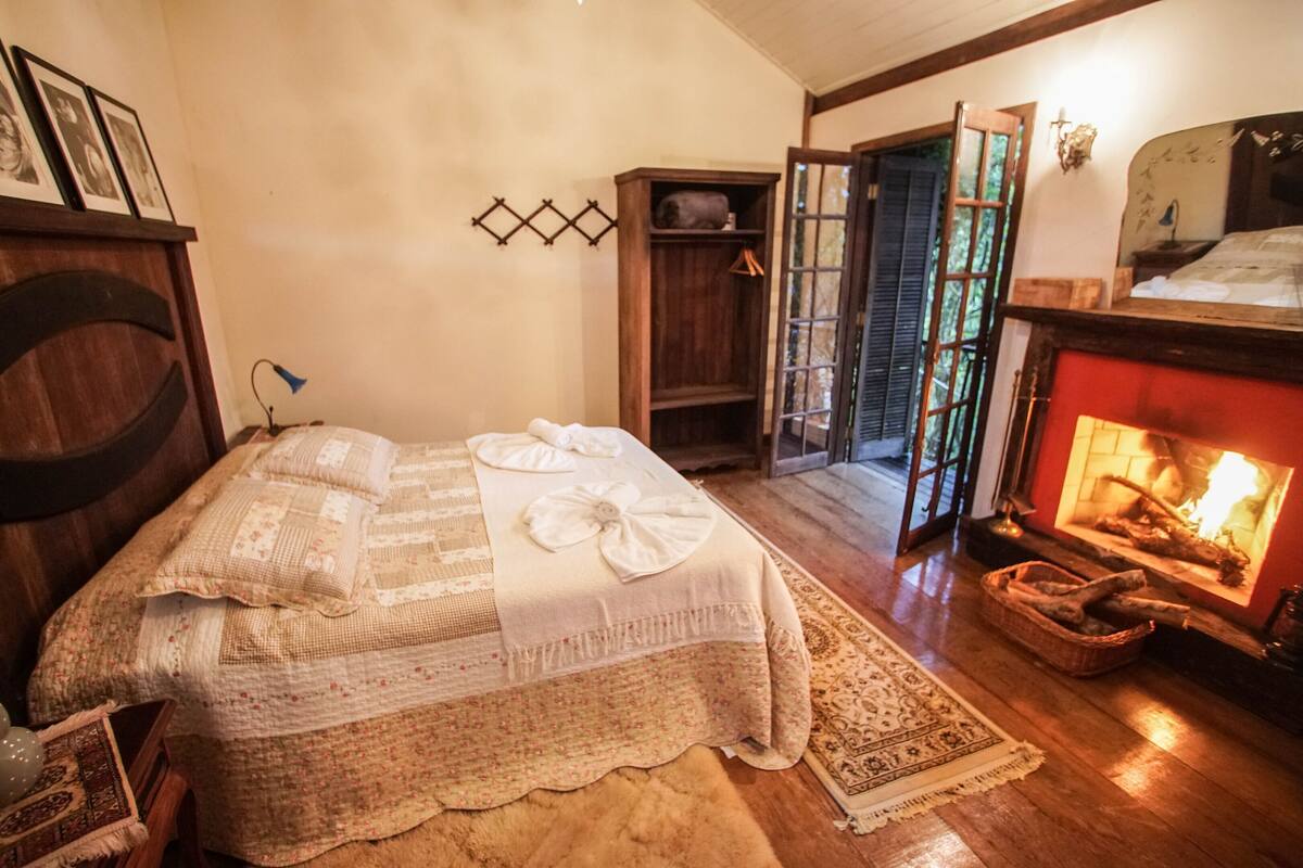 Quarto com cama, lareira, guarda-roupa de madeira e porta que dá para outro espaço.