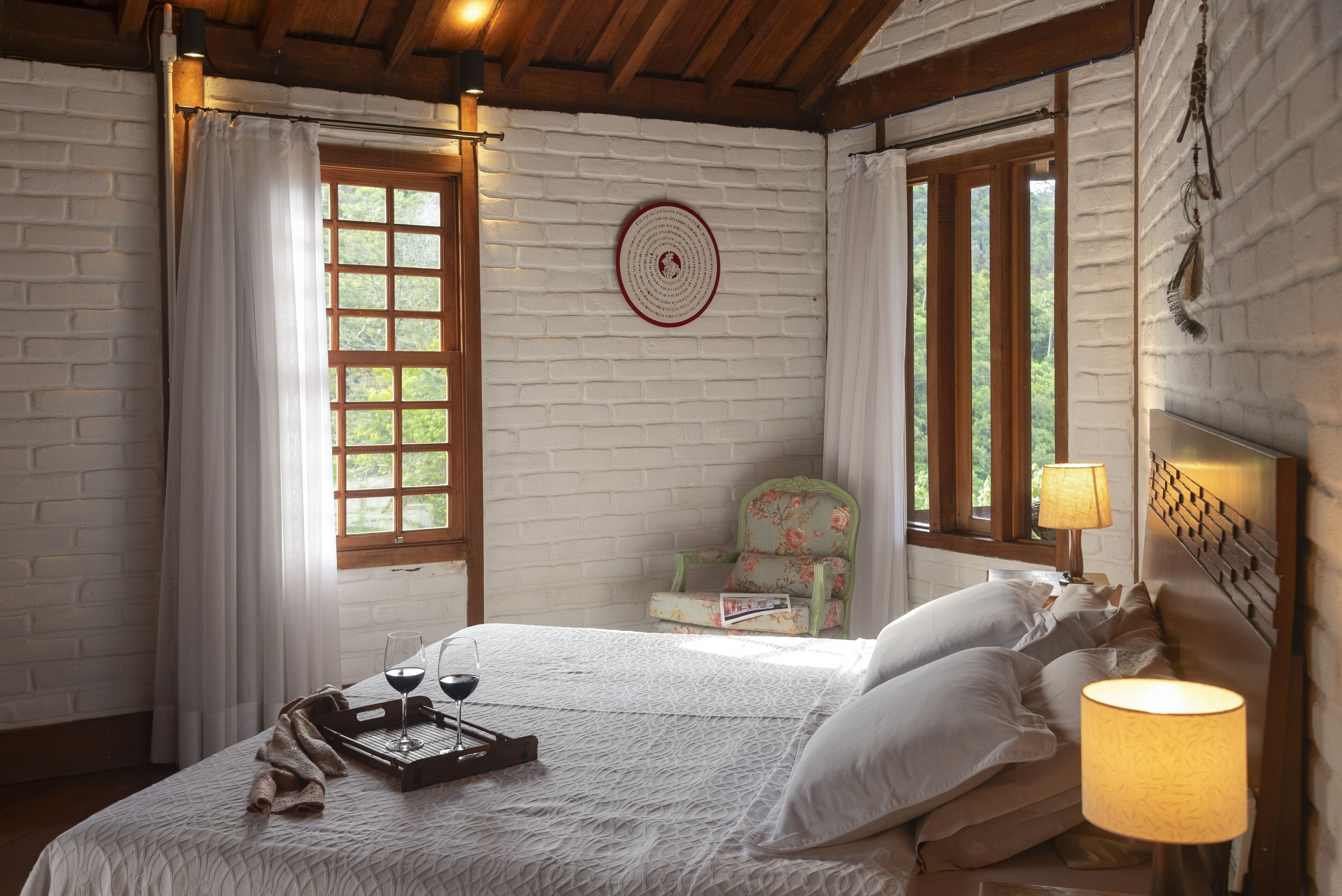 Ein gemütliches Schlafzimmer mit Bett, Nachttisch mit Lampe, einem Fenster mit Vorhängen und einem Sessel.