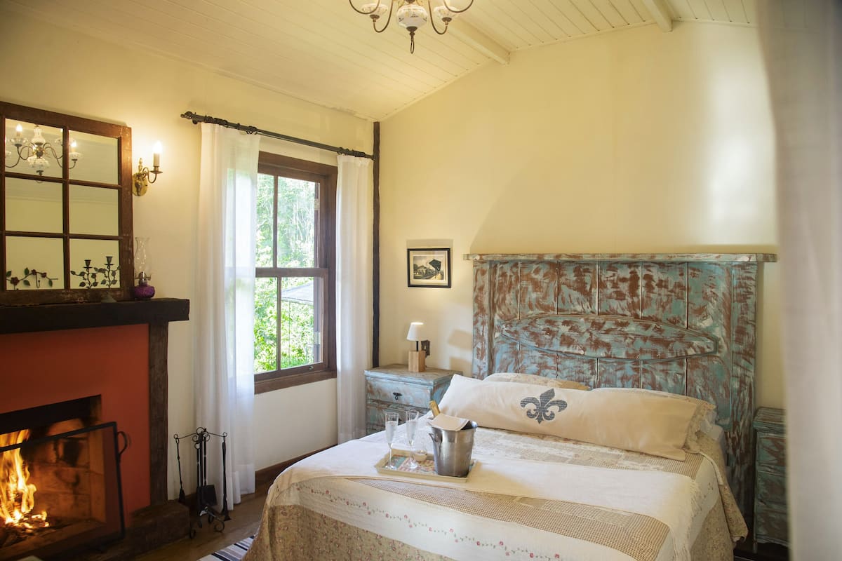 Quarto com lareira, cama com roupa de cama floral, mesa de cabeceira com abajur e janela com cortinas.