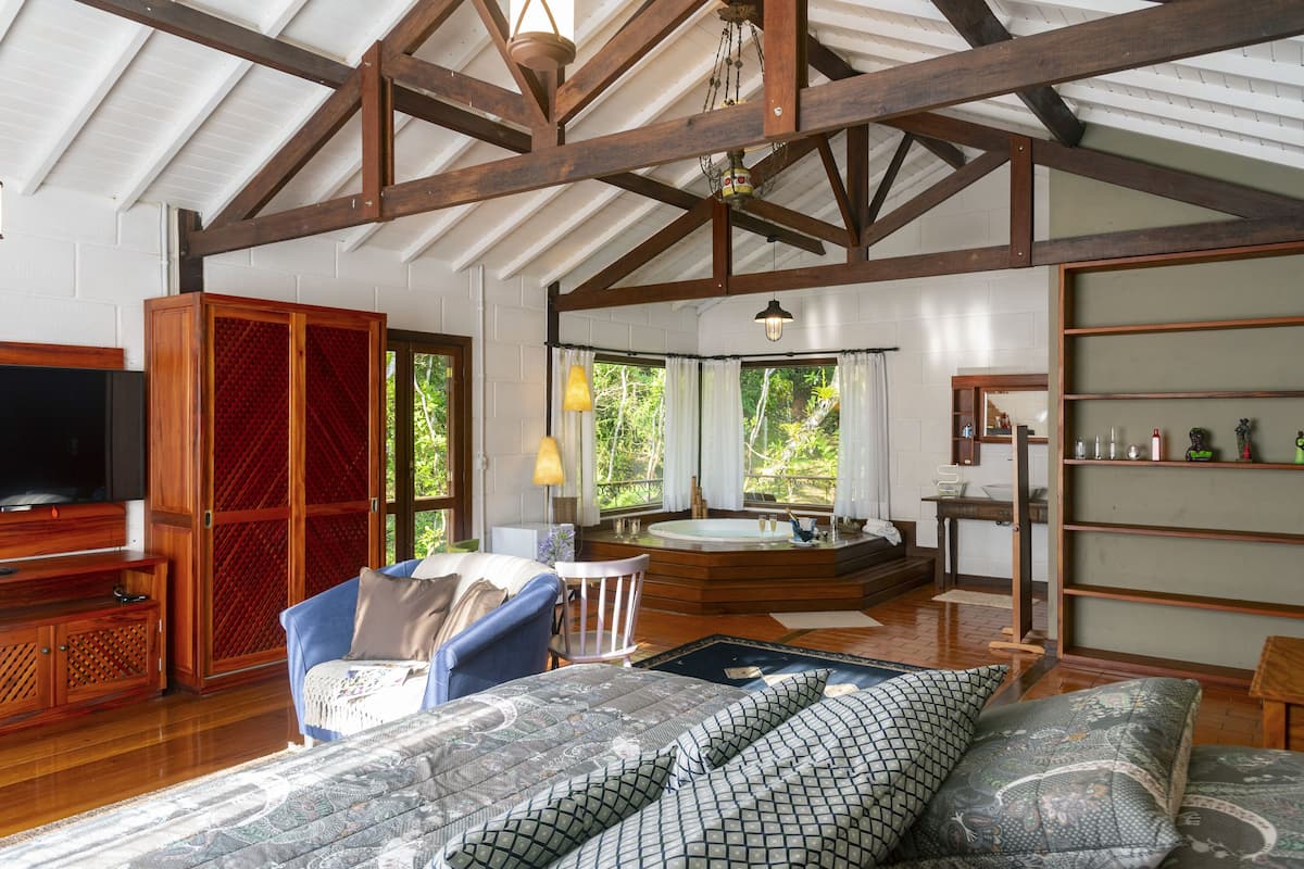 Um quarto amplo com uma cama grande, um rack de madeira para a TV, uma área de estar com uma poltrona, uma mesinha e uma banheira.