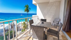 Un balcon avec une table et des chaises, donnant sur une mer d’un bleu limpide, où l’on aperçoit des bateaux et un palmier.