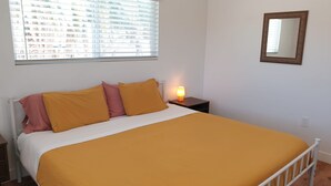 Un dormitorio con cama, mesita de noche con lámpara, ventana con persianas y espejo adosado a la pared.