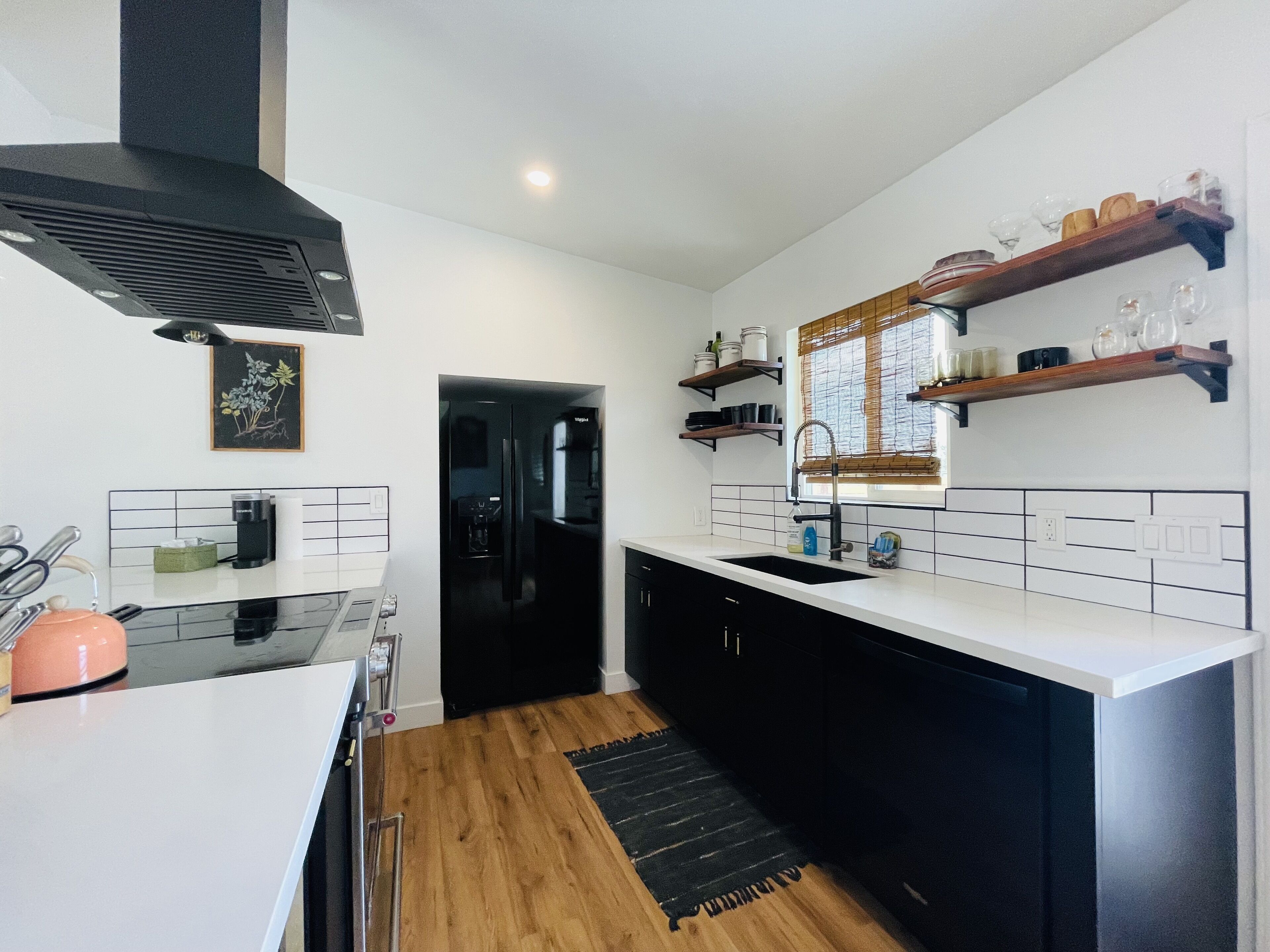 A modern kitchen with a black refrigerator, white countertops, and wooden flooring. There are shelves above the sink holding glassware and jars.