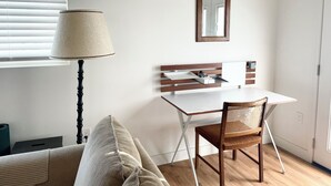 Una habitación pequeña y limpia, con un escritorio blanco, una silla, una lámpara y un espejo en la pared.