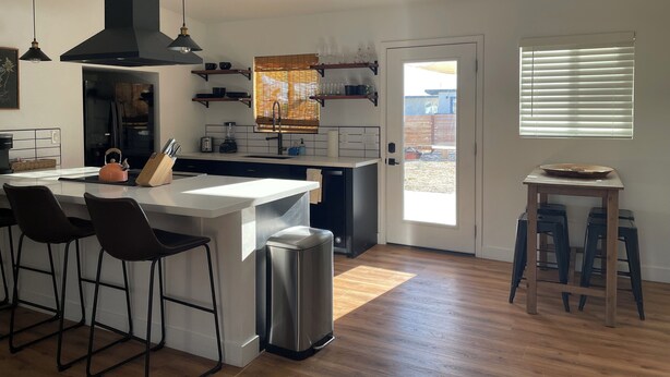 Una cocina moderna con una isla central, taburetes de bar, un bote de basura y vistas al patio trasero por una puerta.