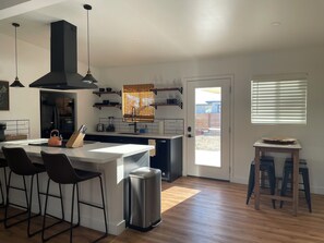 A modern kitchen with a central island, bar stools, a trash can, and a view of the backyard through a door.