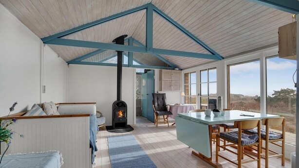 Une pièce chaleureuse avec un plafond en bois, une cheminée, un coin repas avec une table et des chaises, et une vue dégagée grâce à de grandes fenêtres.