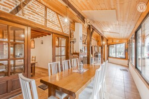 A spacious dining area with a long wooden table and white chairs, brick walls, and large windows providing natural light.