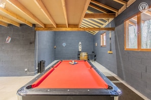 A pool table in a room with dark walls, wooden beams, and a window.