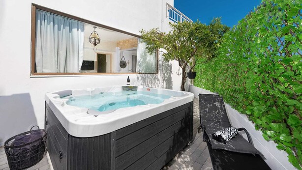 A hot tub outside a white building with a window, a hanging plant, and a chair.