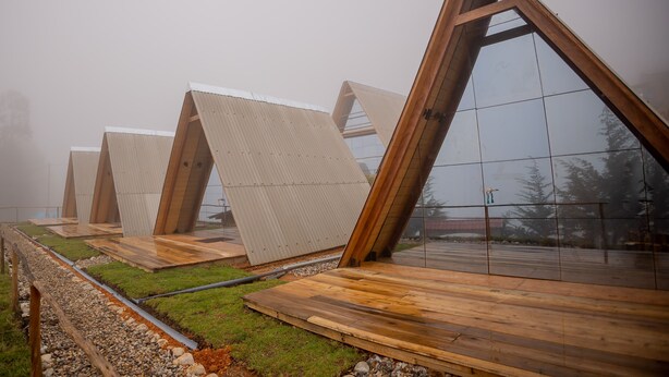 Una hilera de estructuras triangulares de madera con paneles de vidrio reflectante, ubicadas en un césped con un sendero de grava.
