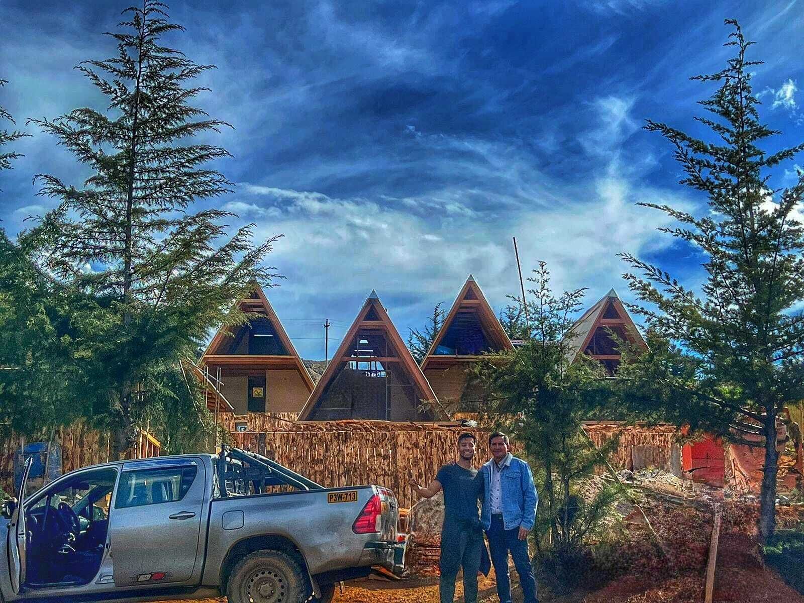 A cabin with a triangular roof design, surrounded by trees, and a parked truck in the foreground.
