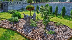 Jardín ajardinado con arbustos variados y un árbol ornamental, delimitado por un muro de piedra y una valla.