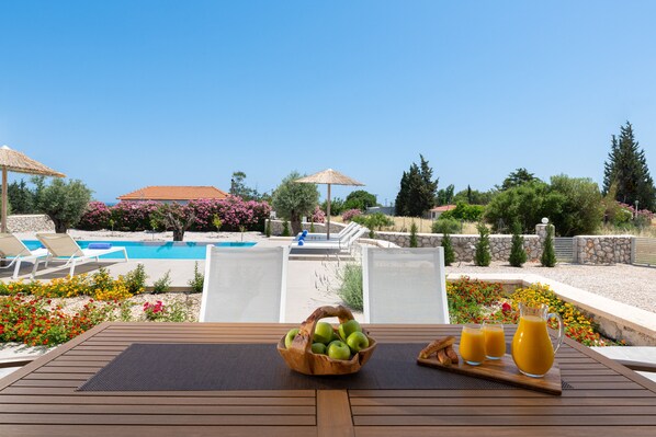 A wooden table with a fruit bowl, juice glasses, and a pitcher, overlooking a pool and garden. - 3 charming newly built semi-detached luxury villas in south rhodes (Γεννάδι)