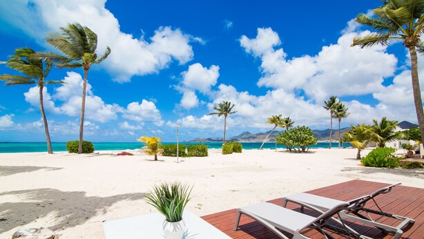 Eine Strandlounge mit weißem Tisch und Stühlen, einer Topfpflanze und Blick auf das Meer und die Berge.