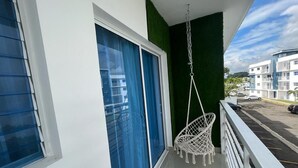 A balcony with a white hanging chair, a blue door, and a view of a parking area and buildings. - Apartamento en moca (Moca)