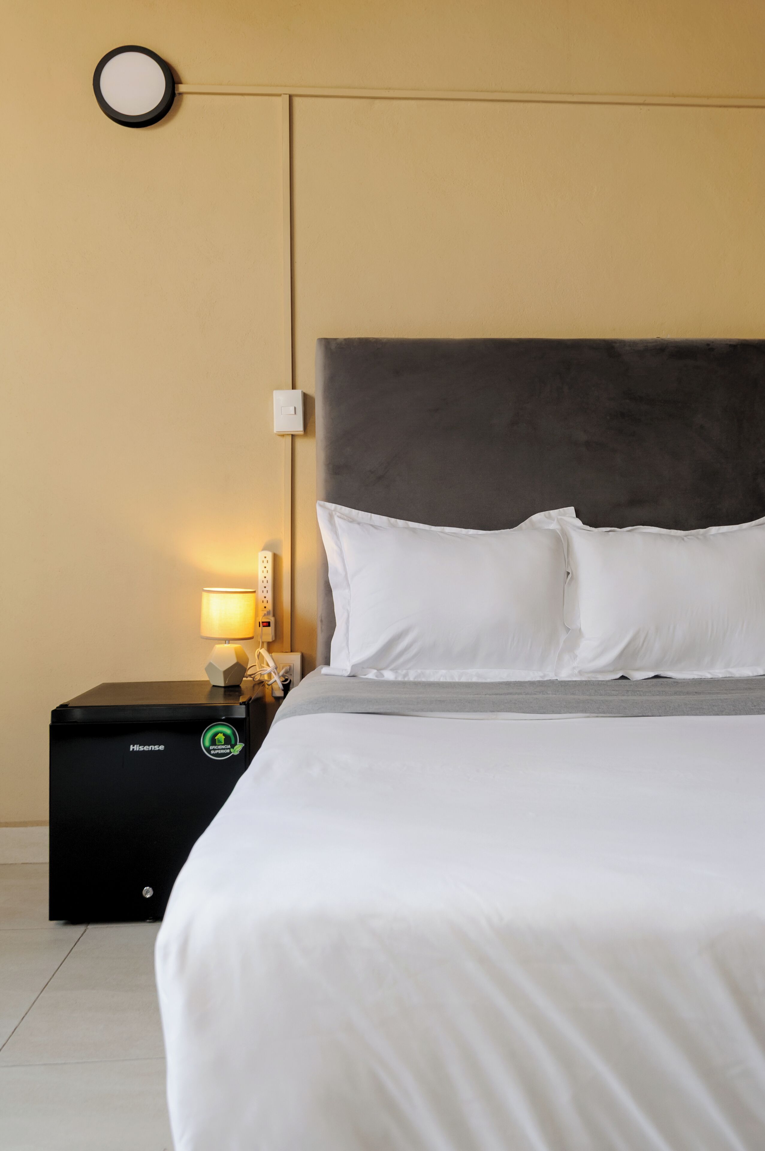 A neatly made bed with white bedding, a grey headboard, and a bedside table with a lamp and a black device labeled 'Hisense.'