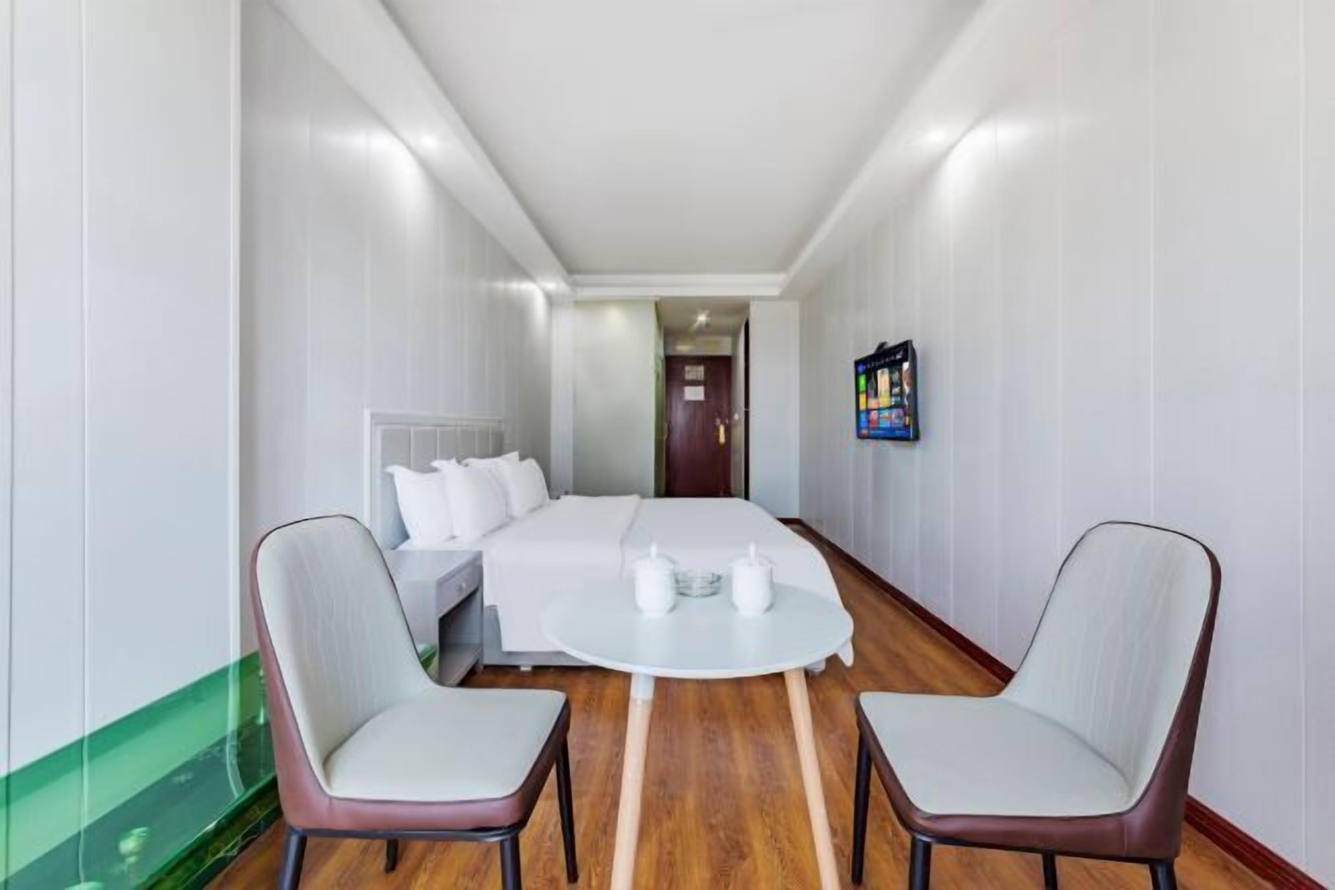 Quarto de hotel com uma cama, duas cadeiras, uma mesinha e uma TV na parede.