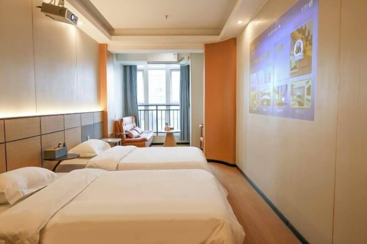 A hotel room with two beds, a desk, a chair, and a large screen displaying images.