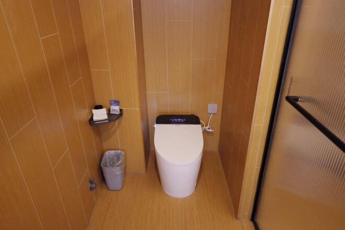 A small, compact bathroom with a toilet, a trash bin, and a shelf with toiletries.