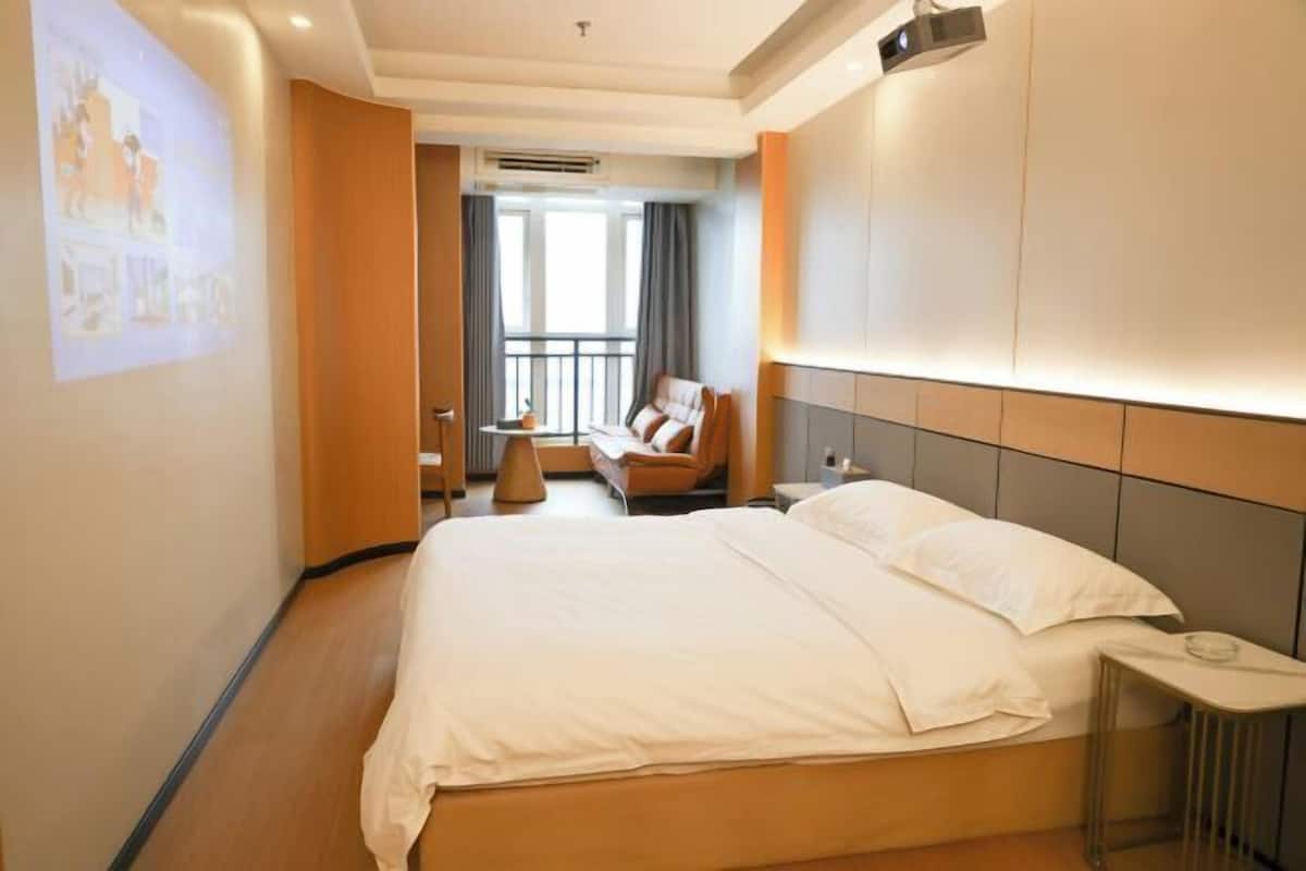 A hotel room with a large bed, a small seating area with a sofa, a side table, and a projector screen on the wall.