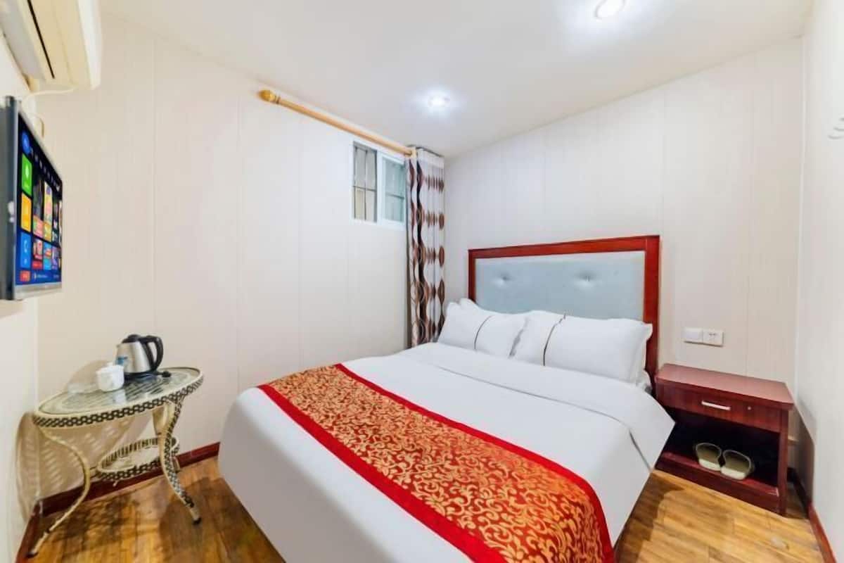 A hotel room with a bed, a small table with a kettle, a nightstand, and a window with curtains.